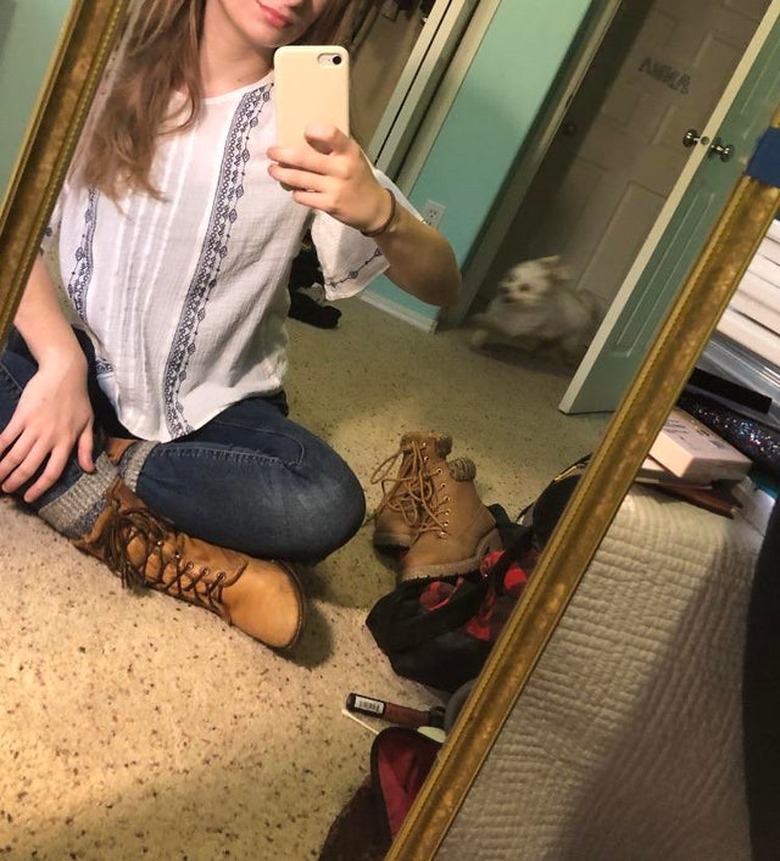 Woman taking selfie in mirror while small white dog runs into the room behind her