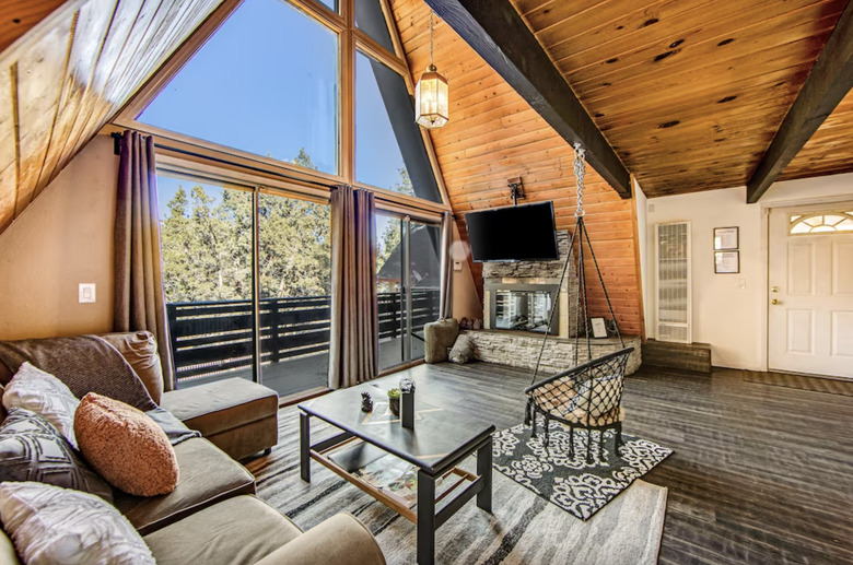 Living room of A-frame with sectional, hanging macrame chair, coffee table, stone fireplace, and a wall-mounted TV.