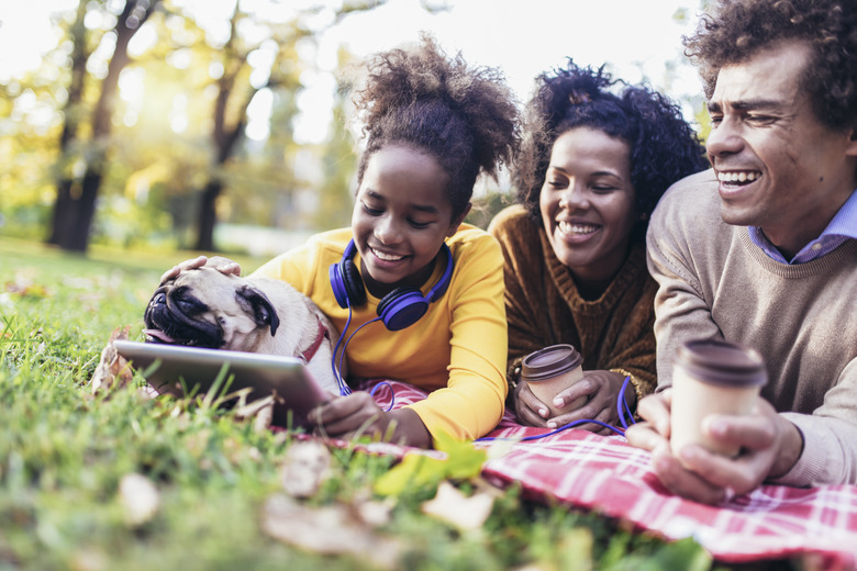 Beautiful young family lying on a picnic blanket with their dog, enjoying an autumn day in park while using digital tablet.