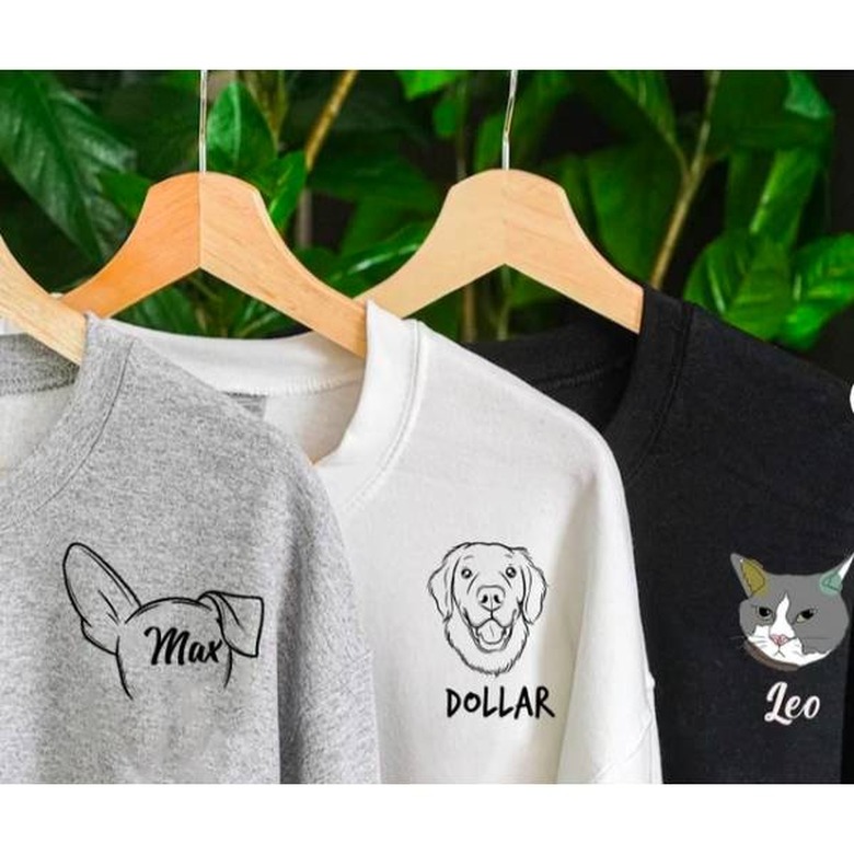 Grey, white, and black shirts with personalized pet illustrations drawn onto them hanging from hangers.