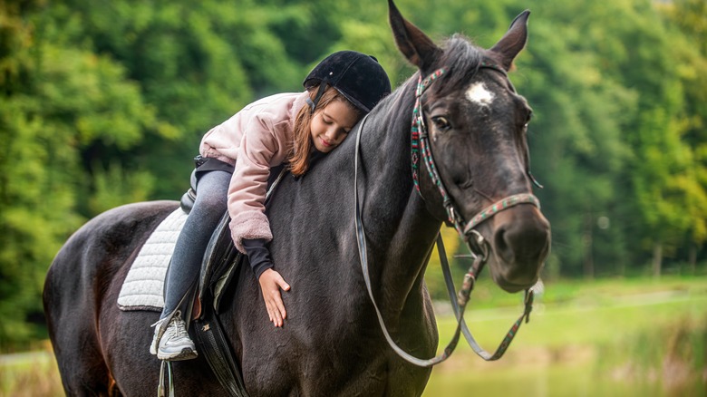 A young girl hugs her black horse while riding it in the forest