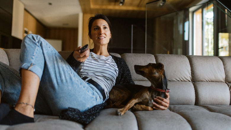 A woman holding a TV remote relaxes on a sofa with a dog