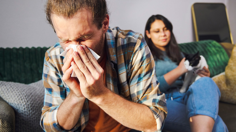 man sneezing into tissue with woman and cat behind him