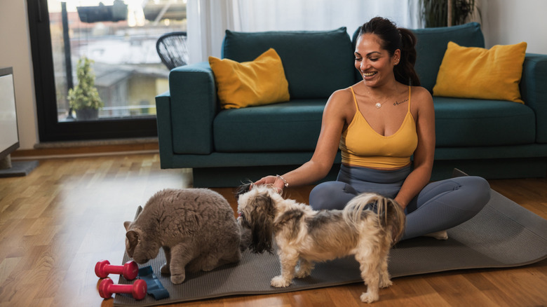 A woman sits on a yoga mat in the living room with her dog and cat