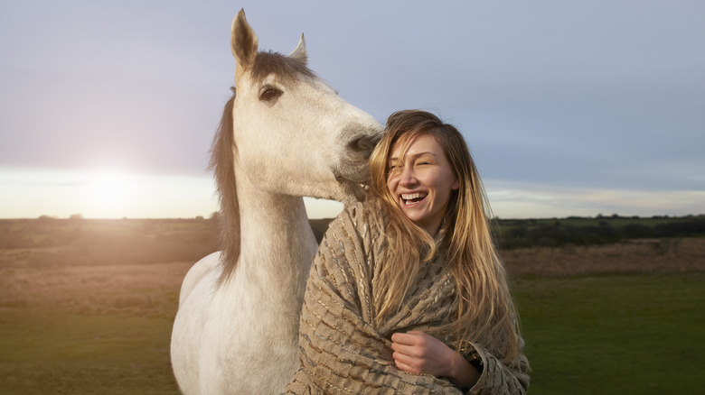 A white horse nibbles on the hair of a blond woman while out in a field