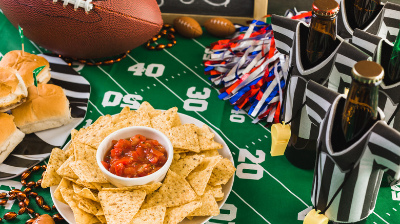 A table of Super Bowl decor and snacks, including bottles with referee shirts, a football field table, chips and salsa, and cheeseburgers.