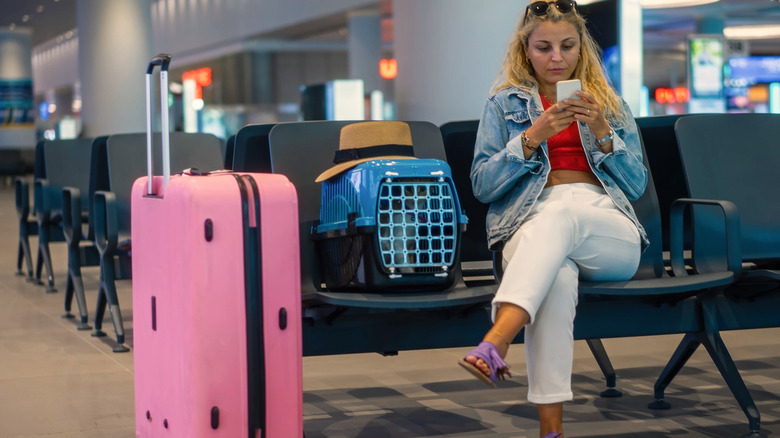 A woman sits at the airport alongside her pet in a carrier and her suitcase