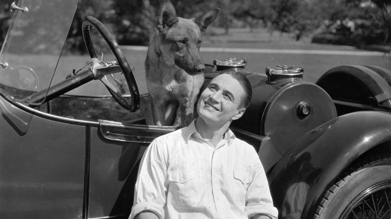 A smiling early 1900s man leans against a car and looks up at a dog