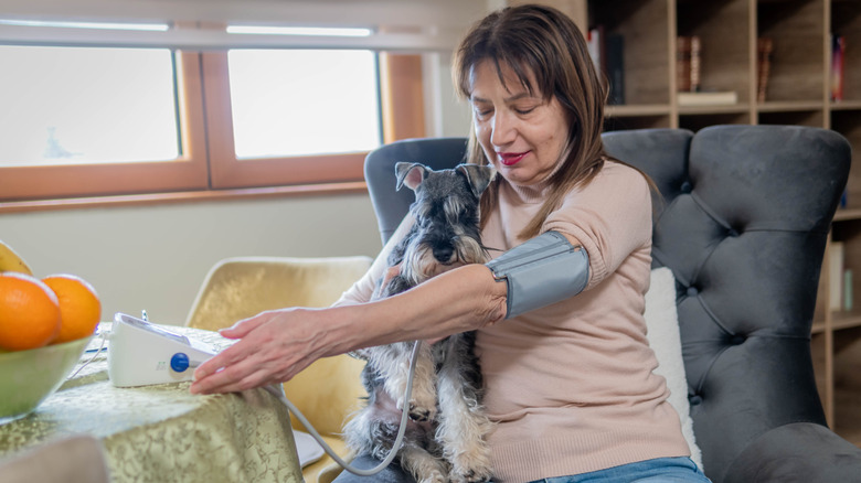 a mature woman takes her blood pressure at home while sitting in a chair and holding her dog