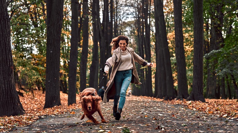 a woman in jeans and a long jacket is running and smiling while holding the leash of a very excited dog