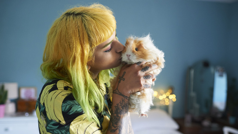 a young person with tattoos and bright yellow and green hair holds a guinea pig and kisses it