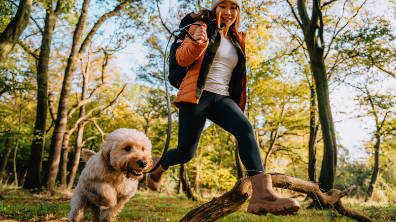 a woman and her golden doodle are running through the forst, jumping over a piece of fallen wood