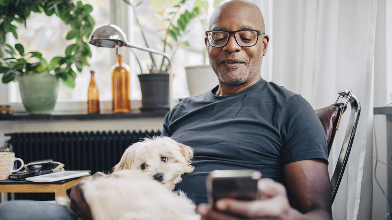 Smiling senior male using smart phone with dog sitting on his lap at home