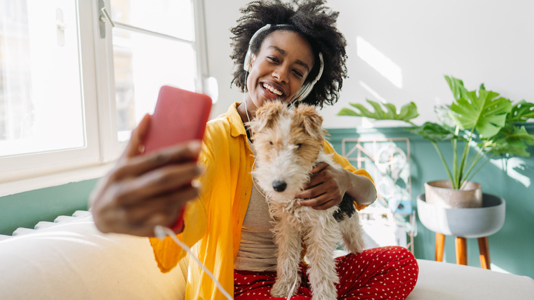 A woman with a dog on her lap is taking a picture inside her bright apartment