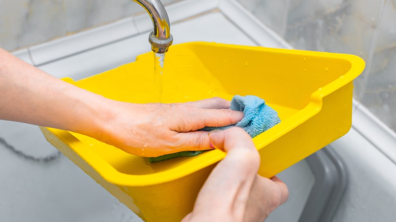 A person scrubs a litter box under a sink with running water.